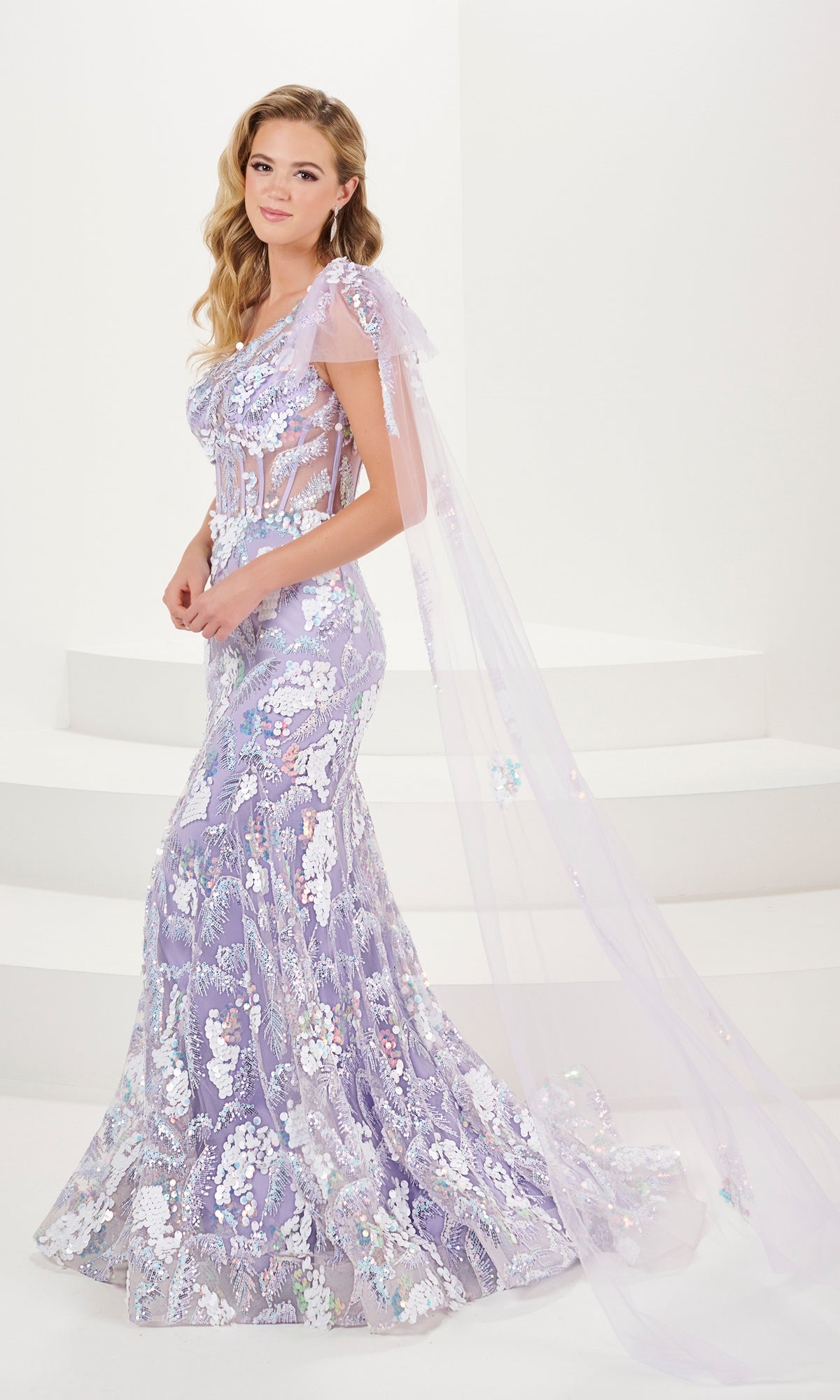 A woman stands on white steps wearing the Tiffany One-Shoulder Sequin Sheer Prom Dress 16082, a fitted lavender and silver mermaid gown featuring sheer panels, a sheer corset bodice, and a long, sheer cape draping from her shoulders.